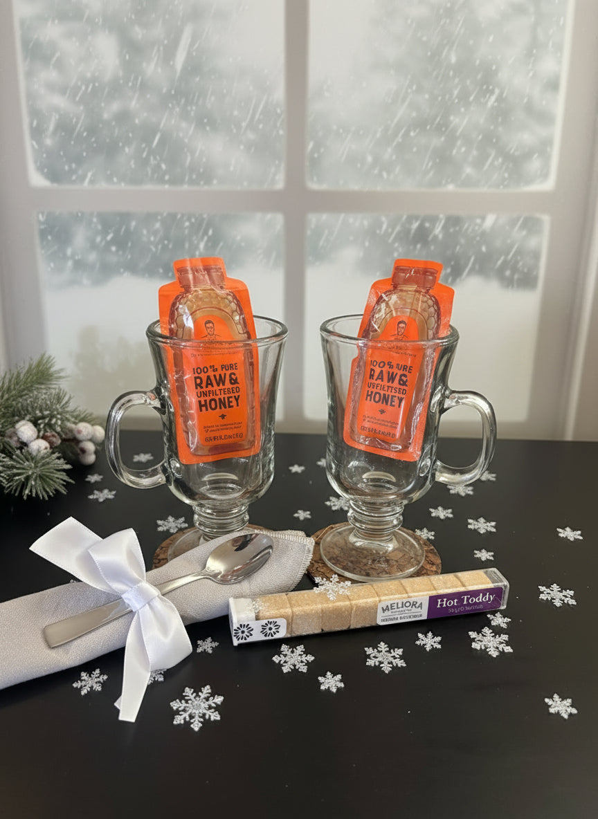 Two glass mugs with hot toddy packets on a table with snowflake decorations and a snowy window background.