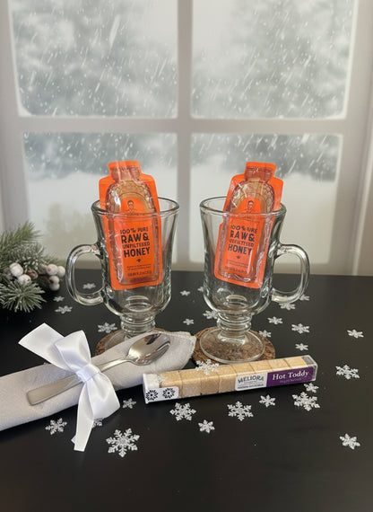 Two glass mugs with hot toddy packets on a table with snowflake decorations and a snowy window background.