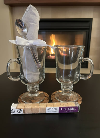 Two glass mugs with a spoon and napkin on top, placed on a book with a fireplace in the background.