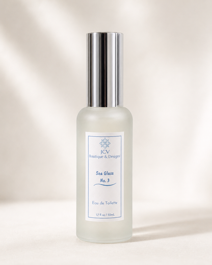 A frosted glass perfume bottle with a shiny silver cylindrical cap and a white label reading 'JCW Boutique & Design Sea Glass No. 3', set against a warm beige background with soft diagonal shadows.