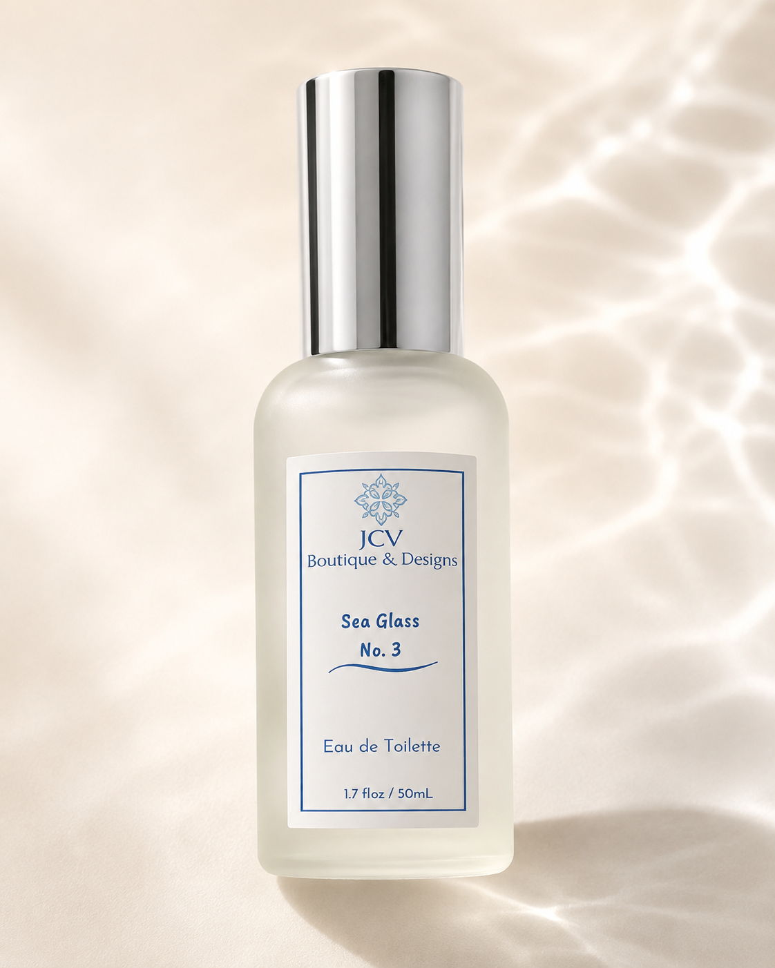 A frosted glass perfume bottle with a reflective silver cap, featuring a white label with blue text that reads 'JCV Boutique & Designs Sea Glass No. 3 Eau de Toilette'.