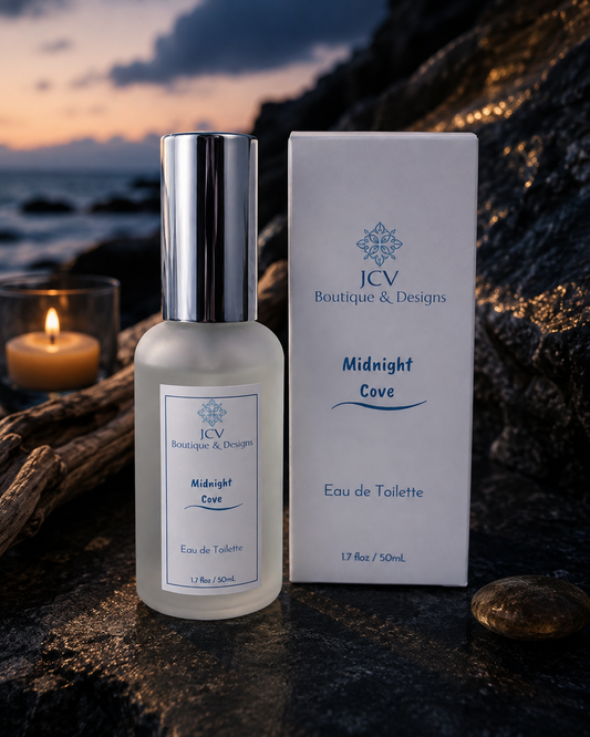 A bottle of JCV Boutique & Designs Midnight Cove Eau de Toilette stands next to its white packaging box on a dark rocky surface, with a lit candle and a sunset ocean scene in the background.